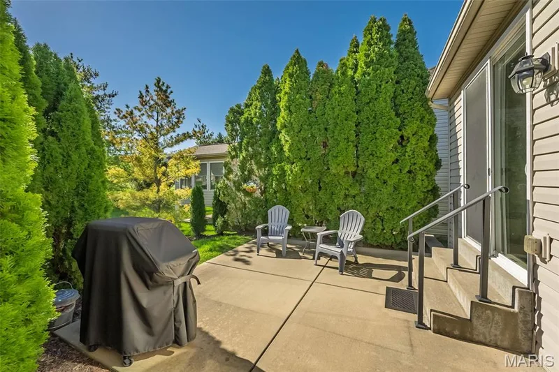 Private patio with a sliding glass door entry, concrete steps, and metal handrails, bordered by mature evergreen trees — 1509 Woodside Hills Drive, St Peters, MO 63376