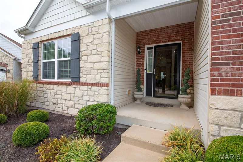 Welcoming entry featuring a covered front porch with stone and brick exterior detailing, black-framed glass door, and multi-pane window with black shutters — 149 Amiot Court, Chesterfield, MO 63146