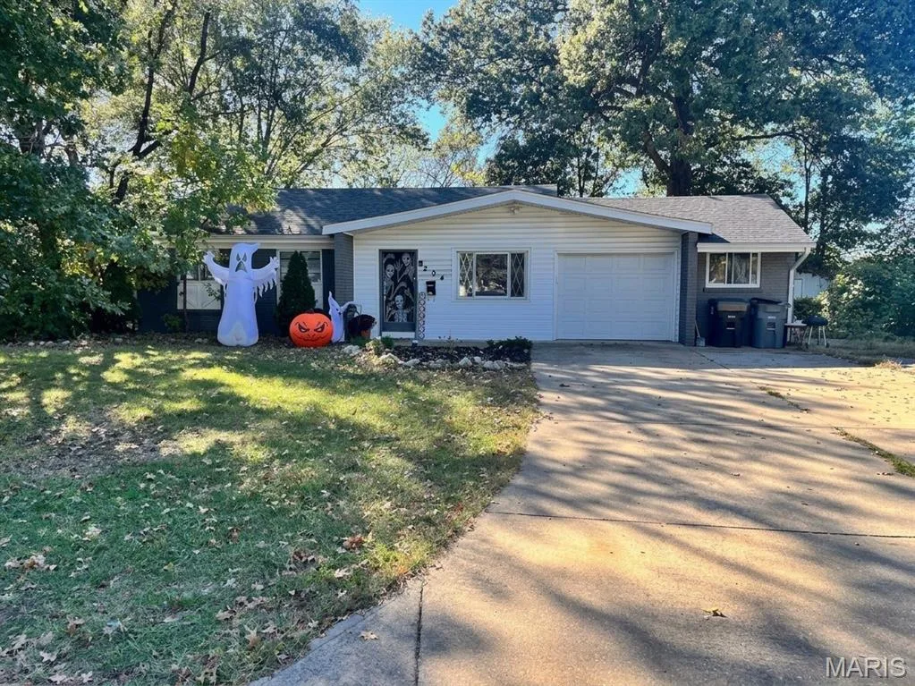 Single-story residence featuring light horizontal siding, a gabled roofline, an integrated one-car garage, and a concrete driveway set against a backdrop of mature trees — 204 Allen Drive, O'Fallon, MO 63366