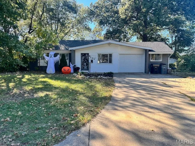 Single-story residence featuring light horizontal siding, a gabled roofline, an integrated one-car garage, and a concrete driveway set against a backdrop of mature trees — 204 Allen Drive, O'Fallon, MO 63366