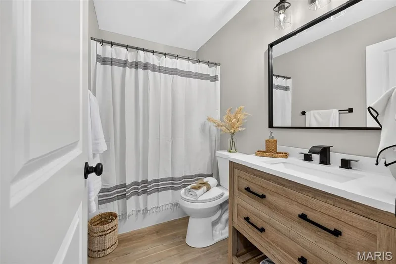 Wood-finish flooring, single vanity with a white countertop, black-framed mirror, and matte black fixtures — 341 Westbrook Drive, O'Fallon, MO 63366