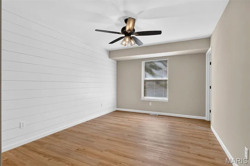 Wood-finish flooring with white baseboards, a ceiling fan with integrated lighting, a window with white trim, a white shiplap accent wall, and a door with black hardware — 341 Westbrook Drive, O'Fallon, MO 63366