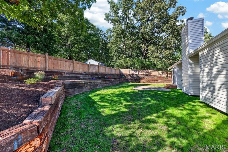 Expansive green lawn with a tiered wood retaining wall, bordered by a privacy fence, and mature trees — 341 Westbrook Drive, O'Fallon, MO 63366