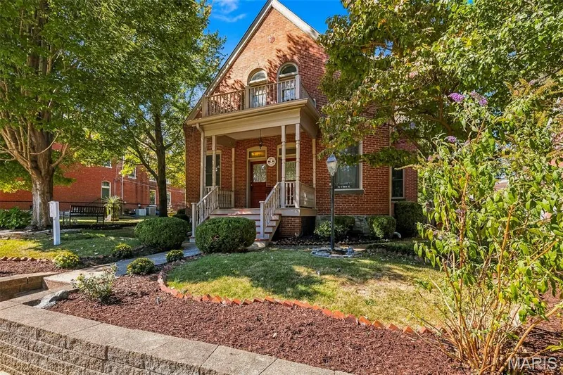 Two-story brick exterior featuring a covered front porch, upper-level balcony with metal railing, arched window detailing, and terraced landscaping with stone retaining walls — 711 N 5th Street, St Charles, MO 63301