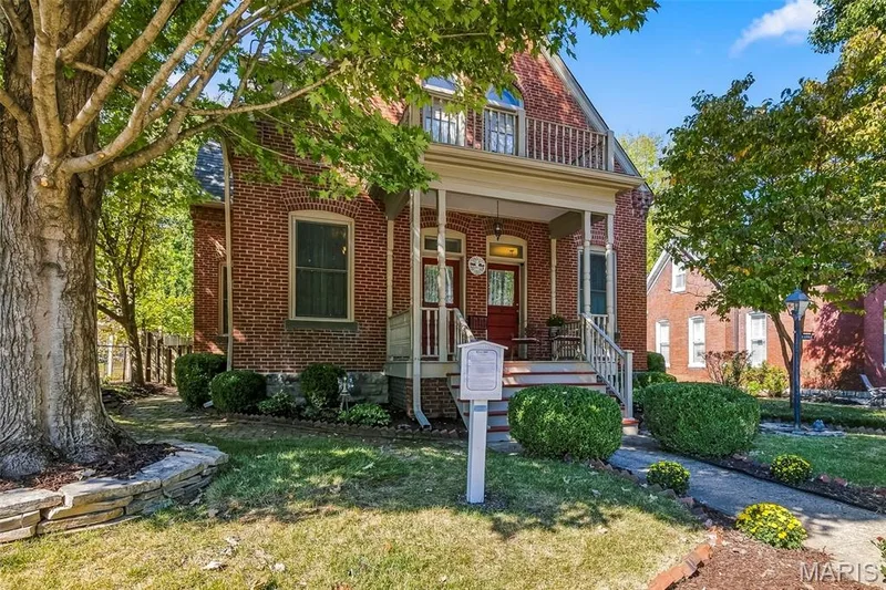 Classic brick exterior featuring a covered front porch, an upper-level balcony with a railing, and stone-bordered landscaping — 711 N 5th Street, St Charles, MO 63301
