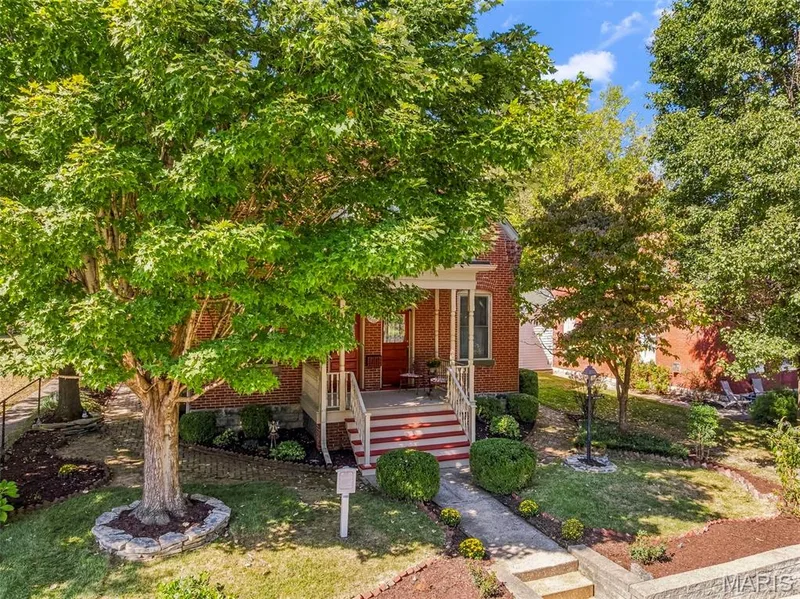 Red brick exterior featuring a covered front porch with ornate columns, concrete walkway, stone-edged garden beds, and mature landscaping — 711 N 5th Street, St Charles, MO 63301