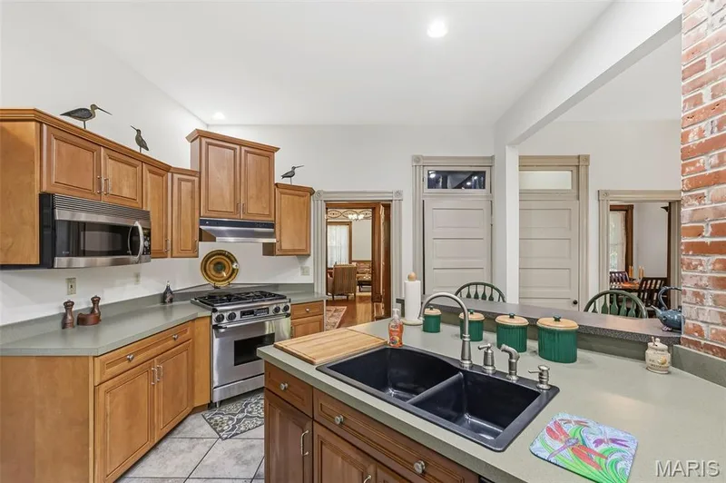 Kitchen featuring wood cabinetry, solid surface countertops, stainless steel appliances, double-basin drop-in sink, and exposed brick column — 711 N 5th Street, St Charles, MO 63301