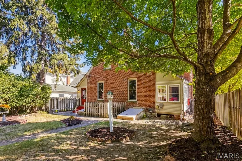 Rear exterior featuring red brick masonry, a wood privacy fence, a basement bulkhead entrance, and mature trees — 711 N 5th Street, St Charles, MO 63301