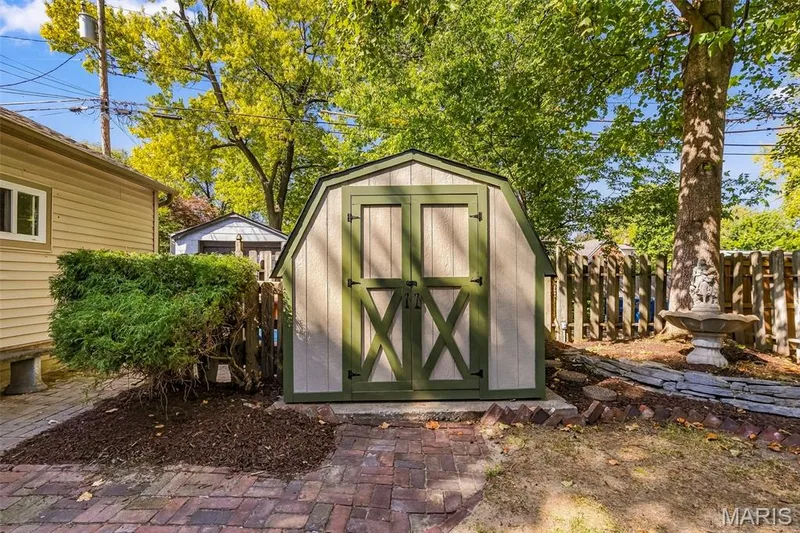 Barn-style storage shed featuring double doors and green trim, set upon a red brick patio with surrounding landscaping, mature trees, and wooden fencing — 711 N 5th Street, St Charles, MO 63301