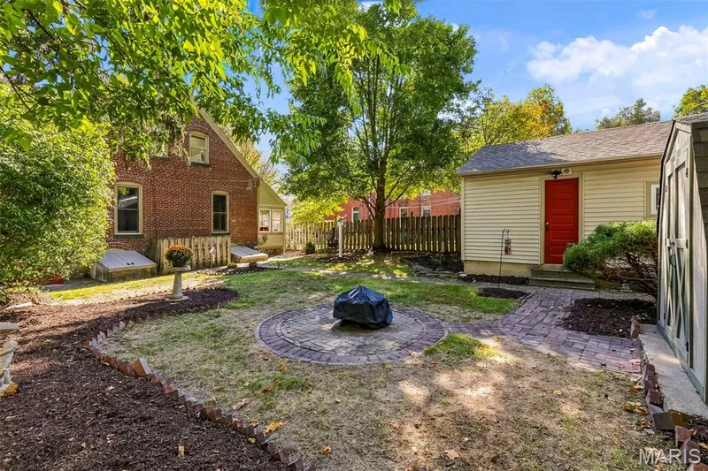 Private backyard featuring a circular brick fire pit, brick-paved pathways, mature landscaping, and a detached outbuilding — 711 N 5th Street, St Charles, MO 63301