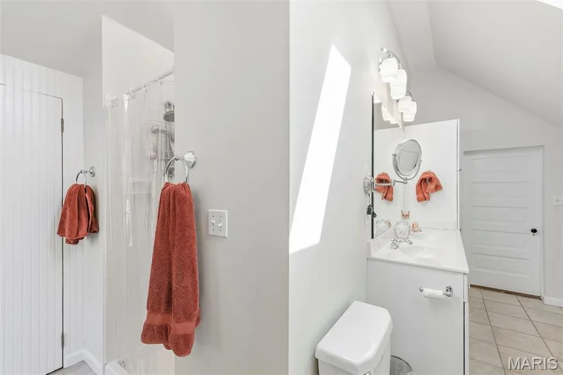 Bathroom featuring a shower stall with a white surround, a vanity with a white countertop and basin, tile flooring, a multi-bulb vanity light fixture, and a wall-mounted magnifying mirror — 711 N 5th Street, St Charles, MO 63301