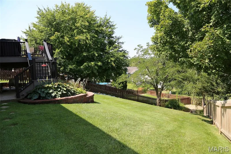 Expansive green lawn with mature trees, a multi-level deck featuring a staircase, a brick-edged planting bed, and a wooden picket fence — 1738 Sun Drop Drive, St Charles, MO 63303