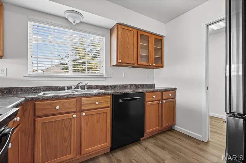 Kitchen featuring wood-finish flooring, wood cabinetry, dark countertops, a double basin sink, and a black dishwasher — 2006 Chapel Hill Crossing, St Peters, MO 63376