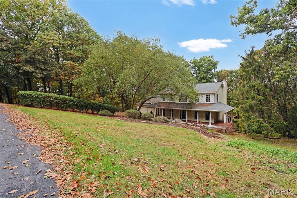 Two-story traditional exterior featuring a covered front porch, light-colored siding, and a steeply pitched roof, set upon a sloped lot surrounded by dense mature trees — 229 Quail Run Drive, Defiance, MO 63341