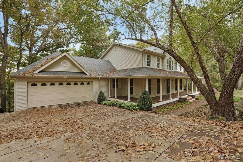 Two-story residence featuring light-toned horizontal siding, a multi-gabled asphalt shingle roof, an attached two-car garage with arched paneling, and a wrap-around porch supported by classical columns — 229 Quail Run Drive, Defiance, MO 63341