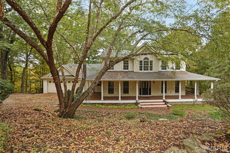 Two-story traditional exterior featuring light-toned siding, a wrap-around porch with column supports, brick porch stairs, and a multi-car garage — 229 Quail Run Drive, Defiance, MO 63341