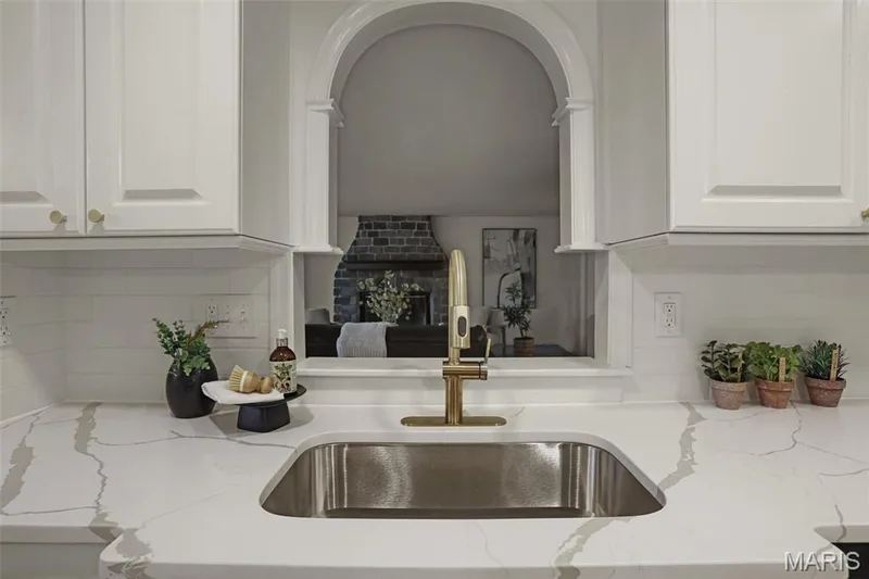 White shaker-style cabinetry, arched pass-through window with architectural detailing, stainless steel undermount sink, metallic finish pull-down faucet, and light-toned countertop with veining — 229 Quail Run Drive, Defiance, MO 63341