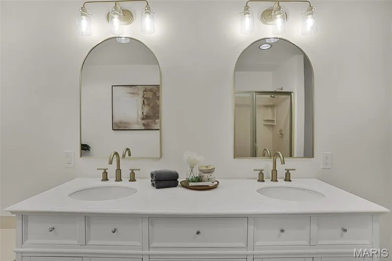 Dual-sink vanity featuring a stone countertop, arched mirrors with gold-tone frames, and three-light brass fixtures — 229 Quail Run Drive, Defiance, MO 63341