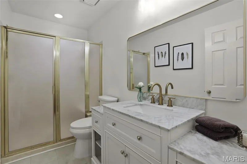 White vanity featuring an integrated stone-composite countertop, undermount sink, brass-finish faucet, and an expansive wall-mounted mirror, paired with a walk-in shower enclosed by a gold-toned frame — 229 Quail Run Drive, Defiance, MO 63341