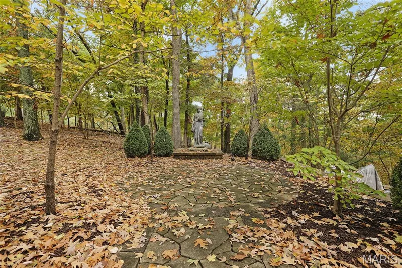 Flagstone patio featuring a stone pedestal statue, manicured shrubbery, and mature hardwood trees — 229 Quail Run Drive, Defiance, MO 63341