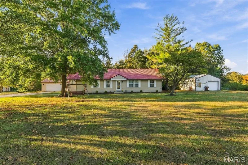 Single-story residence featuring a red metal roof, light-toned siding, attached garage, detached workshop, and expansive grounds with mature trees — 10341 Rose Lane, Foristell, MO 63348
