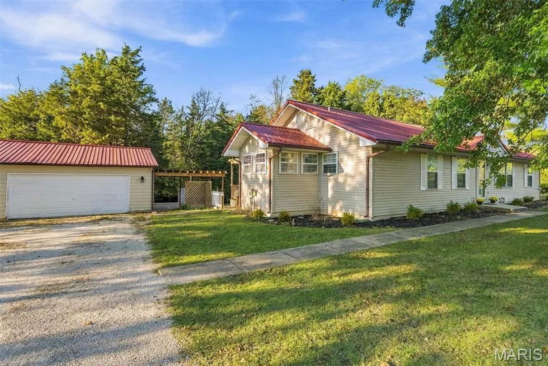 Exterior featuring a neutral-toned siding, a distinct red metal roof, a detached two-car garage, and a timber-framed pergola structure — 10341 Rose Lane, Foristell, MO 63348