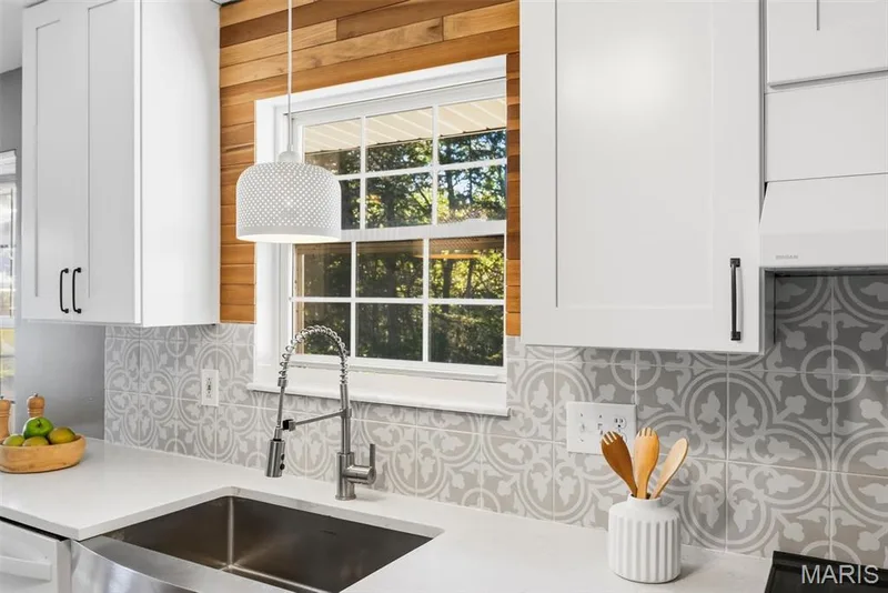 Kitchen featuring shaker-style white cabinetry, patterned tile backsplash, solid-surface countertops, wood-finish wall accent, and a stainless steel farmhouse sink with a pull-down sprayer faucet — 10341 Rose Lane, Foristell, MO 63348