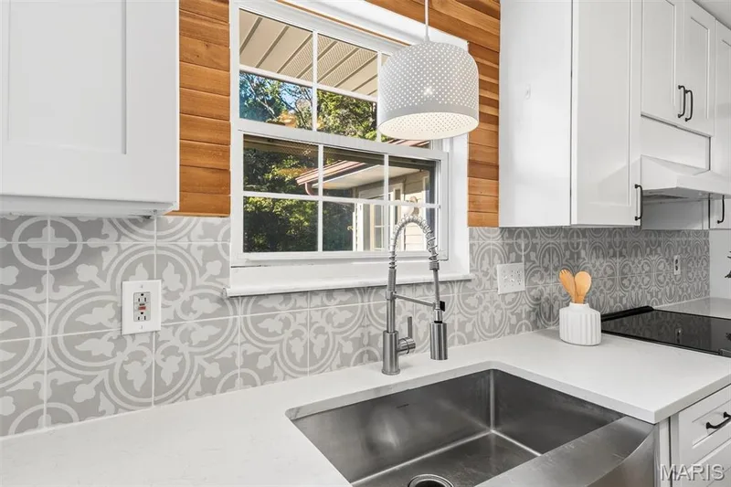 Stainless steel farmhouse sink featuring a high-arc pull-down faucet, white solid-surface countertops, patterned tile backsplash, white shaker-style cabinetry, and horizontal wood-plank wall detailing — 10341 Rose Lane, Foristell, MO 63348