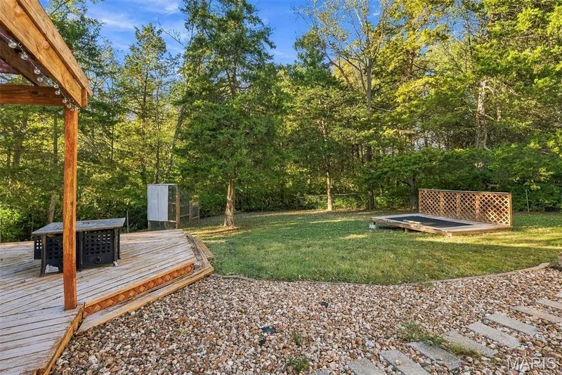 Multi-level wooden deck featuring a lattice railing, stone-surfaced ground cover, manicured lawn, and natural wooded perimeter — 10341 Rose Lane, Foristell, MO 63348