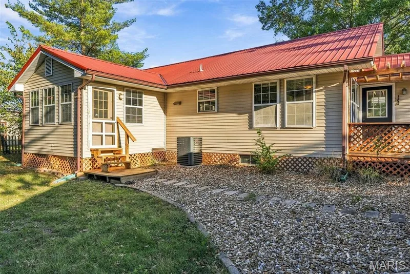 Exterior featuring red metal roofing, light-toned horizontal siding, lattice skirting, a wood deck with stairs, and a stone-lined walkway — 10341 Rose Lane, Foristell, MO 63348