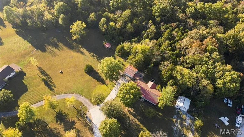 Aerial perspective of a sprawling property featuring expansive cleared pasture, mature tree stands, a gravel driveway, and multiple outbuildings — 10341 Rose Lane, Foristell, MO 63348