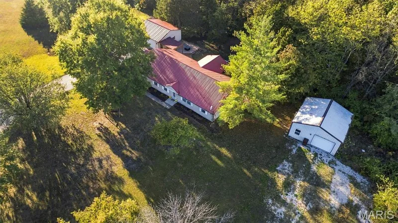 Aerial perspective of the property featuring a residence with a metal roof, an auxiliary outbuilding, a gravel driveway, and surrounding wooded acreage — 10341 Rose Lane, Foristell, MO 63348