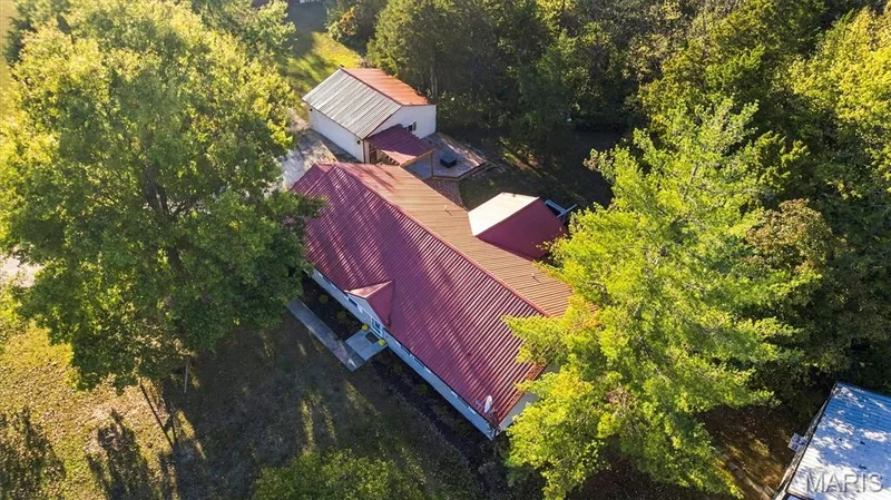 Aerial perspective of property featuring a metal roof, detached garage, hardscaped patio area with wood pergola, and mature trees on a wooded lot — 10341 Rose Lane, Foristell, MO 63348