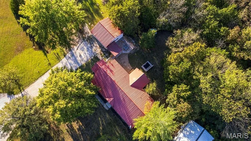 Aerial perspective featuring a metal-roofed residence, detached garage, hardscaped patio area, mature tree canopy, and expansive grass acreage — 10341 Rose Lane, Foristell, MO 63348