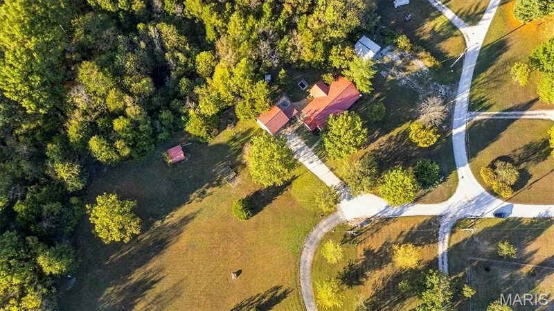 Aerial perspective of property featuring a metal roof structure, paved driveway, expansive grass acreage, and mature tree clusters — 10341 Rose Lane, Foristell, MO 63348