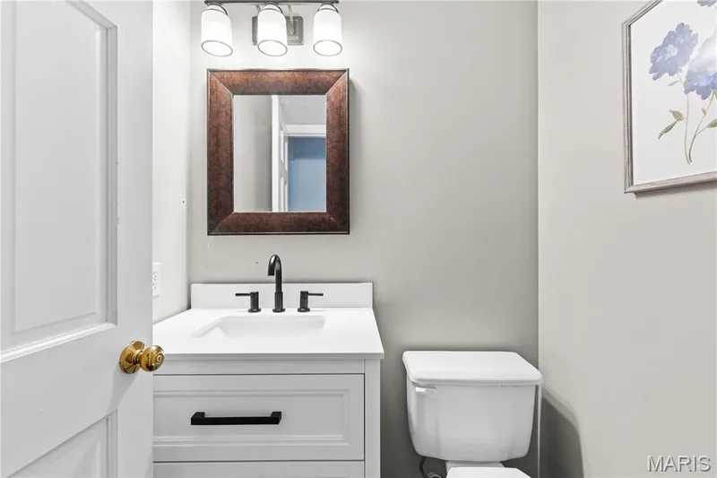 White vanity featuring a rectangular integrated sink, white countertop, and matte black hardware, accented by a three-light vanity fixture and a framed mirror — 838 Emerald Place Drive, St Charles, MO 63304