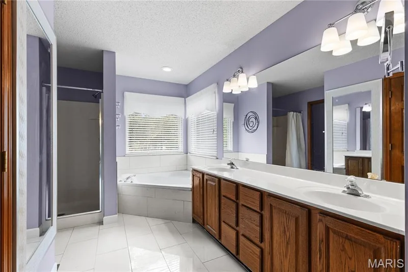 Dual-sink vanity with wood-finish cabinetry and white countertop, corner soaking tub with tiled surround, glass-enclosed shower, and large wall-mounted mirrors — 838 Emerald Place Drive, St Charles, MO 63304
