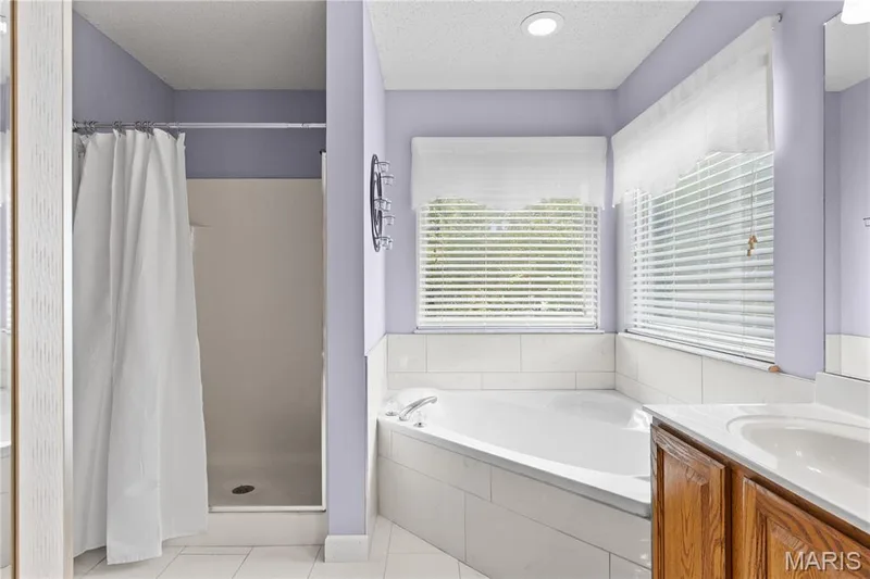 Corner soaking tub with white tile surround, separate shower enclosure, wood-finish vanity with an integrated basin, and horizontal window blinds — 838 Emerald Place Drive, St Charles, MO 63304