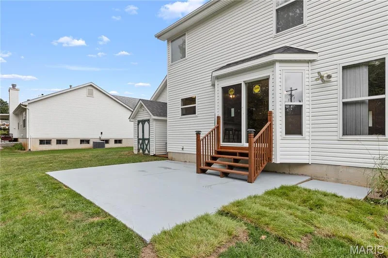 Rear exterior featuring light-colored horizontal siding, concrete patio, wood-railed steps, and an attached storage shed — 838 Emerald Place Drive, St Charles, MO 63304