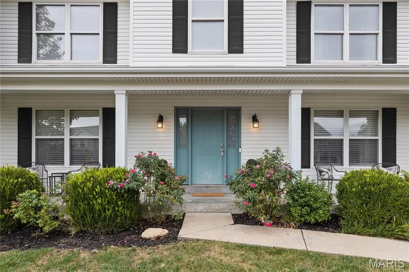 Covered front porch featuring white structural columns, horizontal lap siding, dark shutter accents, and twin exterior wall-mounted light fixtures — 838 Emerald Place Drive, St Charles, MO 63304