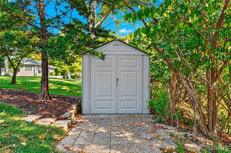 Detached storage structure featuring double doors, situated on a concrete paver pad within a landscaped yard with mature trees — 15552 Chequer Drive, Chesterfield, MO 63017