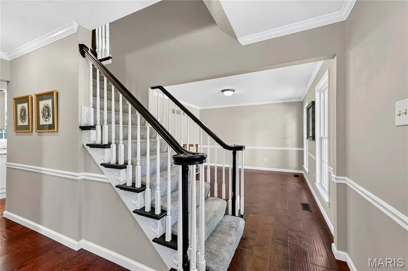 Staircase featuring white balusters, dark-stained handrails, and carpeted treads, complemented by wood-finish flooring and traditional wall molding — 15552 Chequer Drive, Chesterfield, MO 63017
