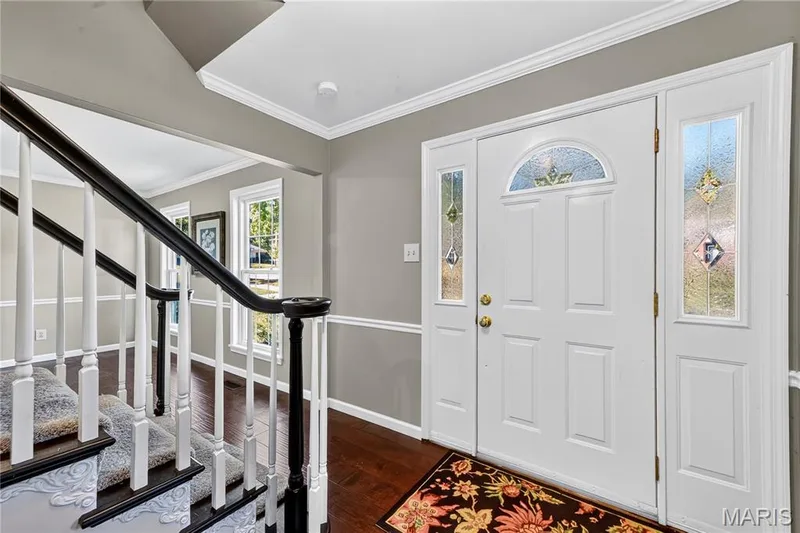 Foyer entrance featuring a white panel door with leaded glass accents and sidelights, wood-finish flooring, and a staircase with a dark-stained handrail and white balusters — 15552 Chequer Drive, Chesterfield, MO 63017
