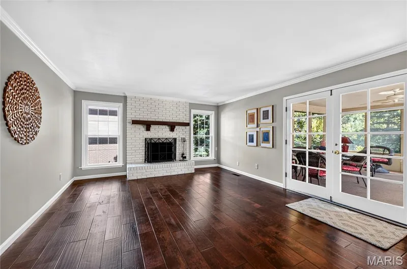 Living space featuring dark wood-finish flooring, white painted brick fireplace with a custom wood mantel, crown molding, and double glass-paned doors — 15552 Chequer Drive, Chesterfield, MO 63017