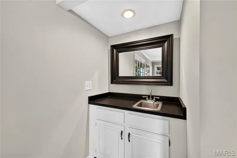 Built-in cabinetry featuring white shaker-style doors, dark solid-surface countertop, recessed stainless steel sink, and framed wall-mounted mirror — 15552 Chequer Drive, Chesterfield, MO 63017