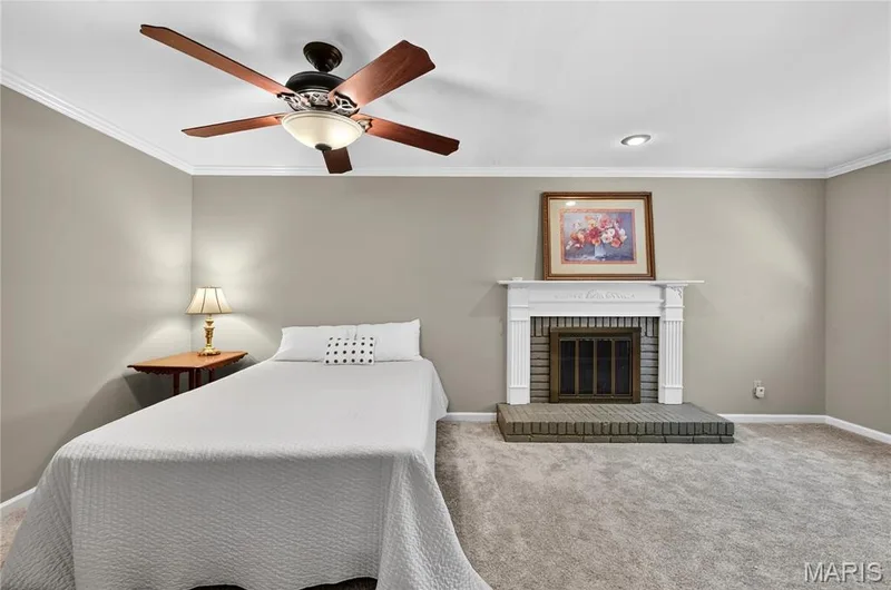 Fireplace featuring a classic white mantel, painted brick surround, and raised brick hearth, complemented by crown molding and a ceiling fan with integrated lighting — 15552 Chequer Drive, Chesterfield, MO 63017