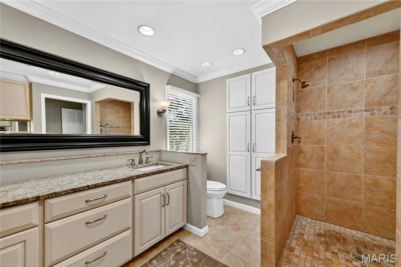 Bathroom featuring a stone-topped vanity with an expansive framed mirror, built-in floor-to-ceiling cabinetry, and a walk-in shower with tiled surround and decorative accent border — 15552 Chequer Drive, Chesterfield, MO 63017