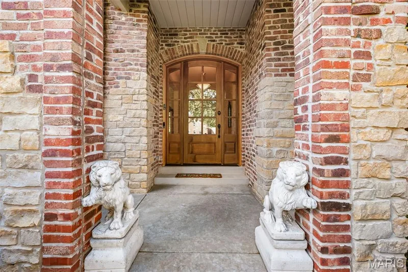 Entryway featuring red brick and natural stone masonry, arched mahogany door with multi-pane glass inserts, and symmetrical stone pedestal sculptures — 112 Castlewood Estates Drive, O'Fallon, MO 63368