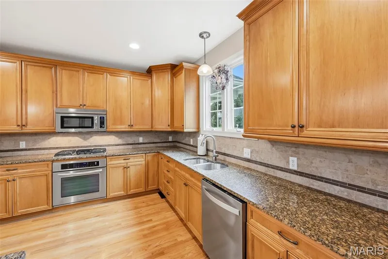 Kitchen featuring natural wood cabinetry, stone-finish countertops, stainless steel appliances, tile backsplash with decorative inlay, and wood-finish flooring — 112 Castlewood Estates Drive, O'Fallon, MO 63368