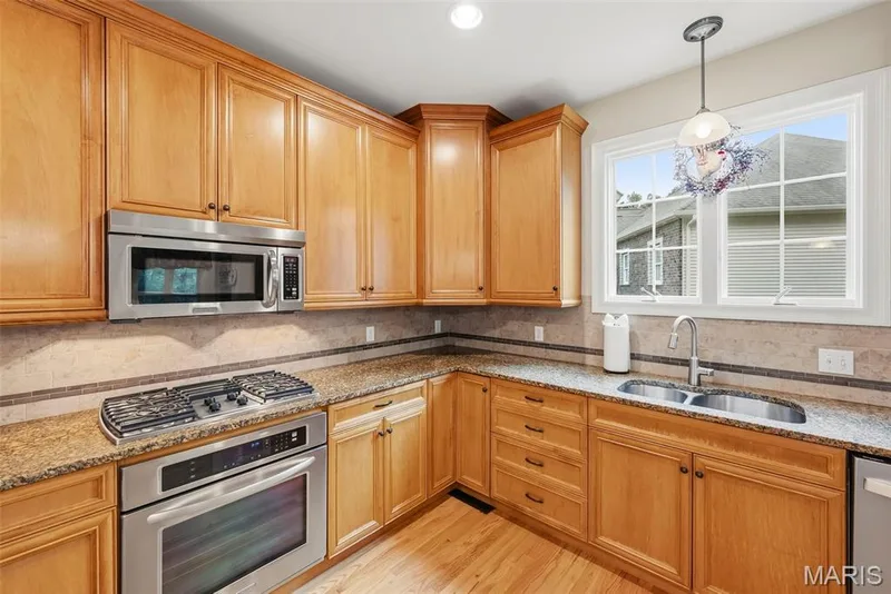 Kitchen featuring natural wood cabinetry, stone countertops, stone tile backsplash with decorative inset, stainless steel gas range with built-in microwave, and stainless steel dual-basin sink with pull-down faucet — 112 Castlewood Estates Drive, O'Fallon, MO 63368